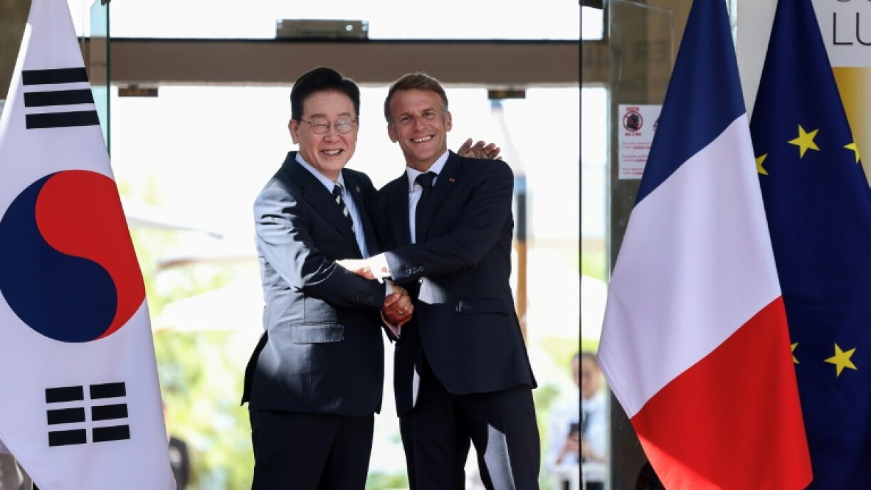 Lee Jae Myung e Emmanuel Macron cumprimentam-se diante das bandeiras da Coreia do Sul, França e União Europeia.